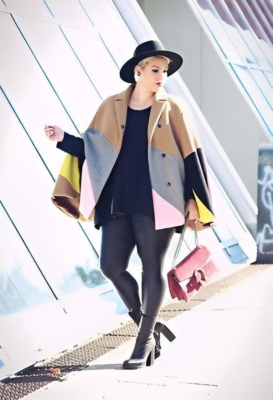 A woman walking outdoors wearing a beige, gray, black, and pink color-blocked cape jacket over a black top and black leggings, with a black wide-brim hat and a pink handbag.