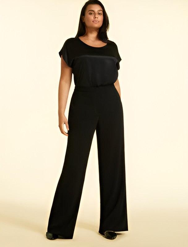 A woman modeling a black jumpsuit with a scoop neckline, short flutter sleeves, and wide-leg pants against a solid cream background.