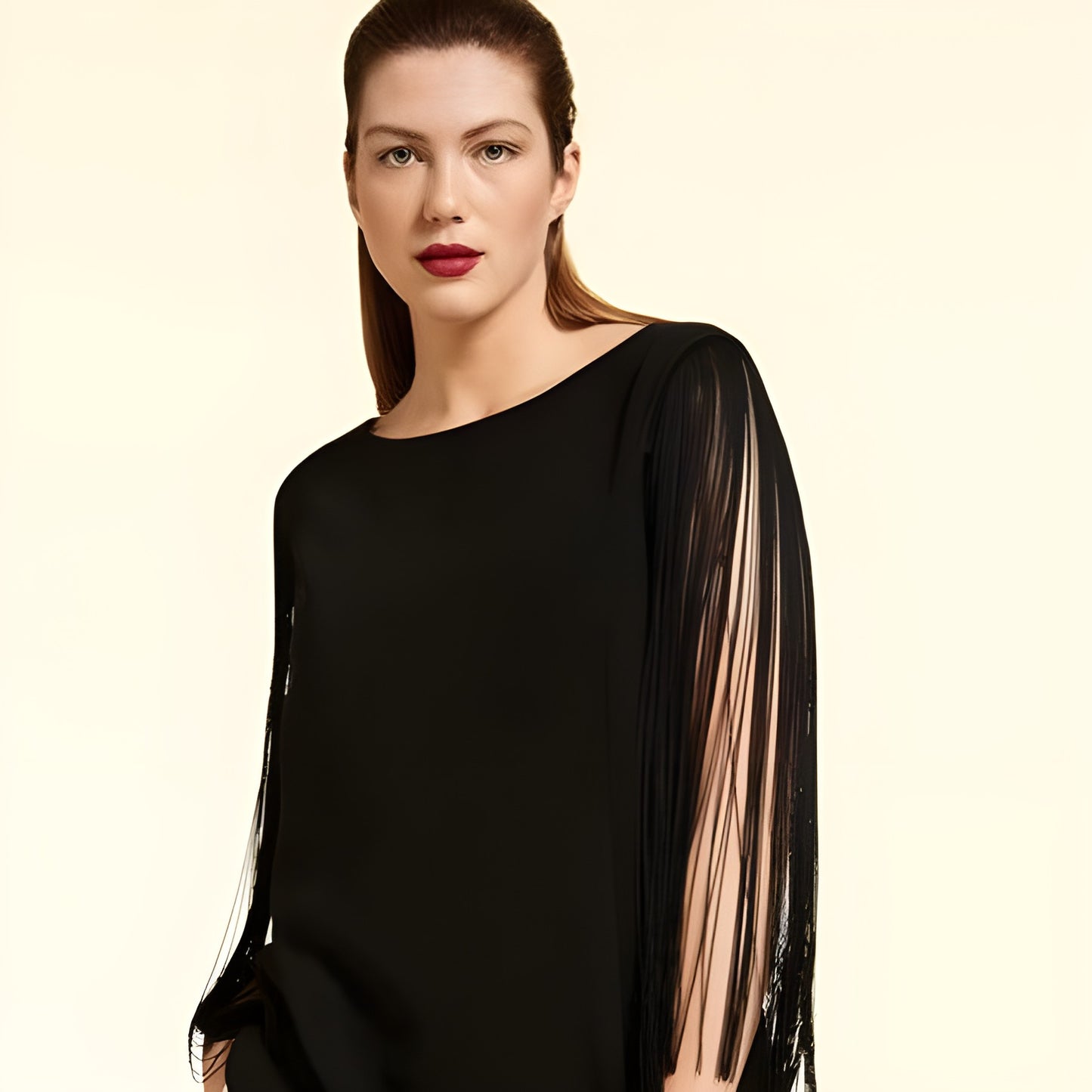A woman modeling a black asymmetrical long-sleeve top with sheer fringe detailing on one sleeve, set against a solid cream background.