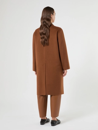 A woman viewed from the back wearing a long brown coat, matching brown trousers, and black shoes against a plain light gray studio background.