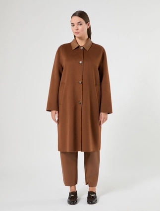 A woman modeling a long brown coat with a pointed collar and front buttons, paired with matching brown pants and black shoes, against a plain light gray studio background.
